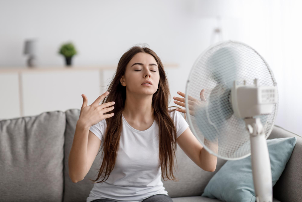 sad-young-european-woman-suffering-from-heat-sits-sofa-catches-cold-air-from-fan-waves-hands