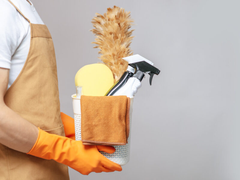 side-view-close-up-hand-young-man-apron-rubber-gloves-holding-basket-cleaning-equipment-feather-duster-spray-bottle-sponge-cloth-wiping-basket